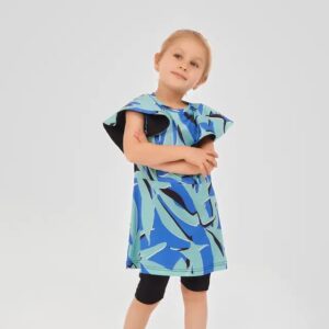 Young girl wearing a two-piece modest swimsuit with a blue and teal abstract leaf print top and black knee-length swim shorts, posing playfully against a light grey background.