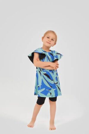 Young girl wearing a two-piece modest swimsuit with a blue and teal abstract leaf print top and black knee-length swim shorts, posing playfully against a light grey background.